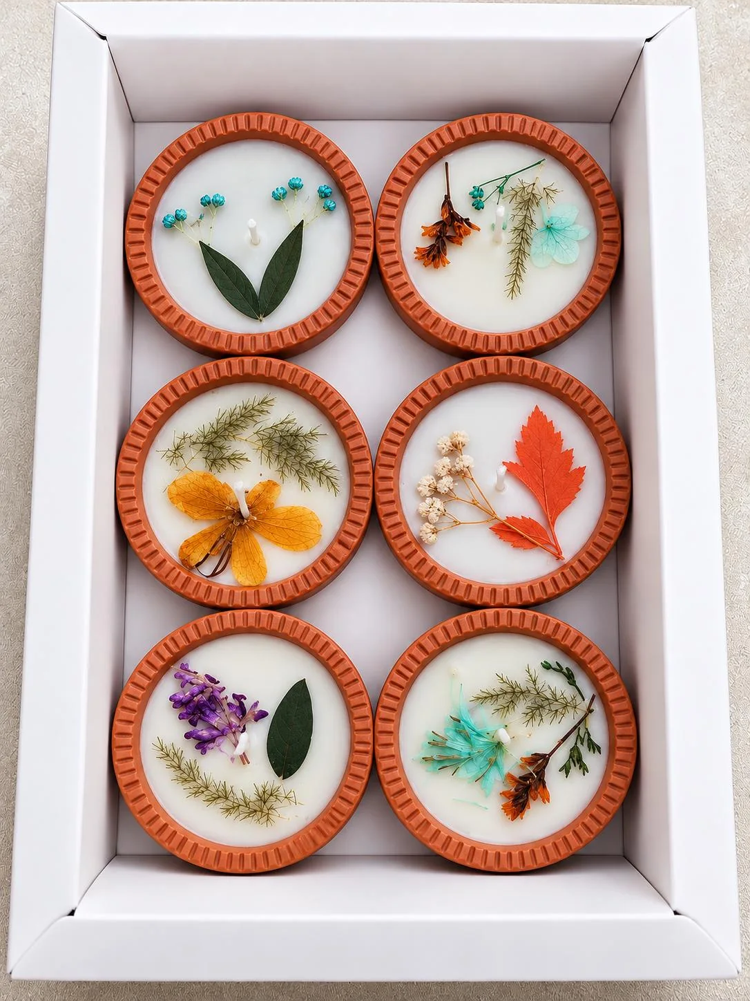 Six terracotta diyas with grooved rims in a white tray, each filled with white wax and decorated with different dried flowers, leaves and fern.